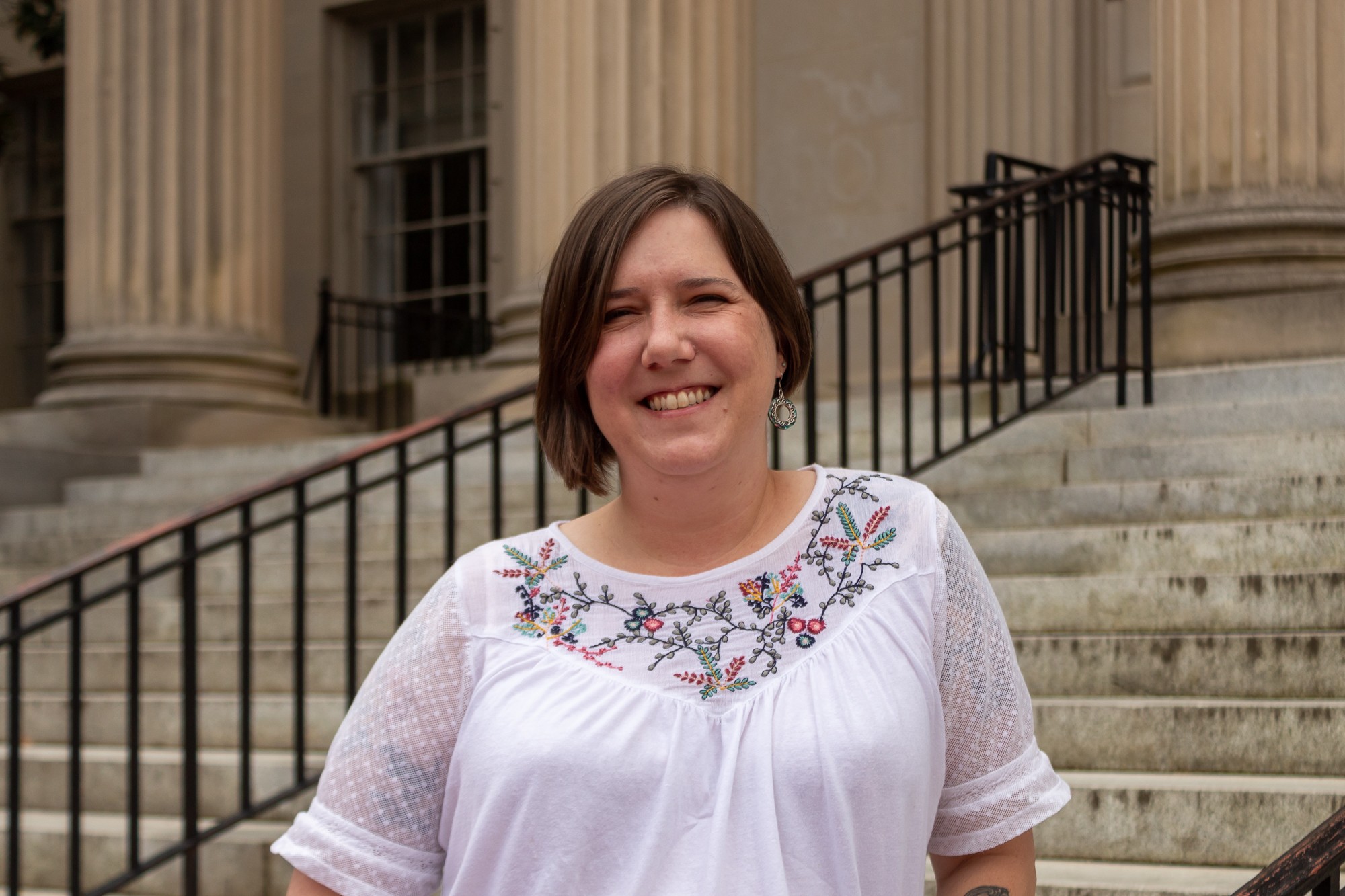 Portrait of Flannery Fitch, a UNC graduate and non-traditional student, smiling on the steps of Wilson Library. She has short brown hair, silver earrings and a white top with embroidered flowers.