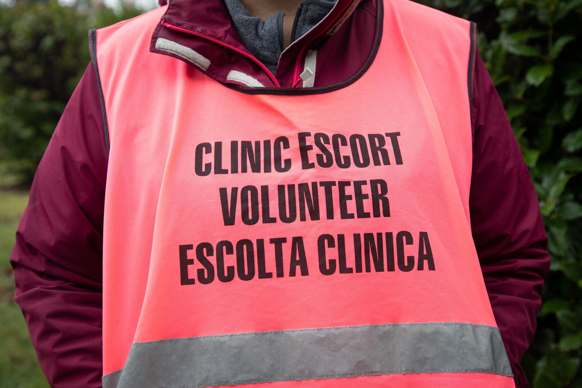 An individual wears a bright pink vest that reads “Clinic Escort, Escolta Clinica.”
