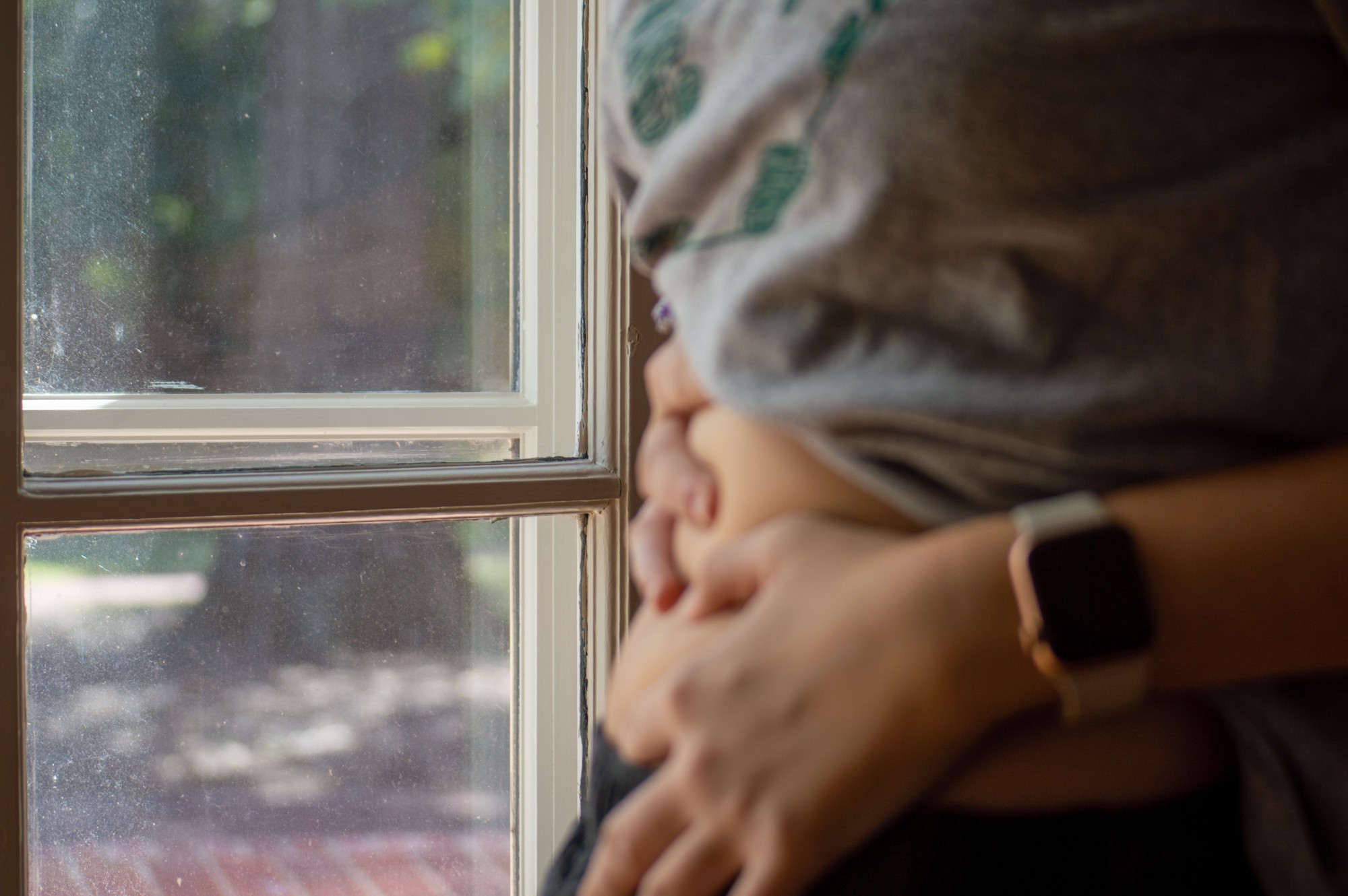 Photo illustration of a person standing by a window, holding their belly as if pregnant. 